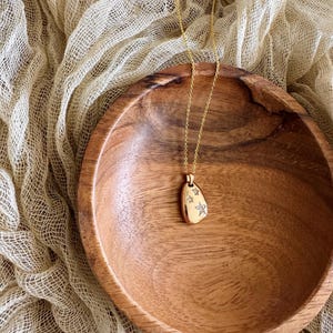 Puede incluir: Un collar de oro con un peque&ntilde;o colgante de forma ovalada que presenta un dise&ntilde;o de estrellas con peque&ntilde;as piedras transparentes. El collar se muestra en un cuenco de madera.