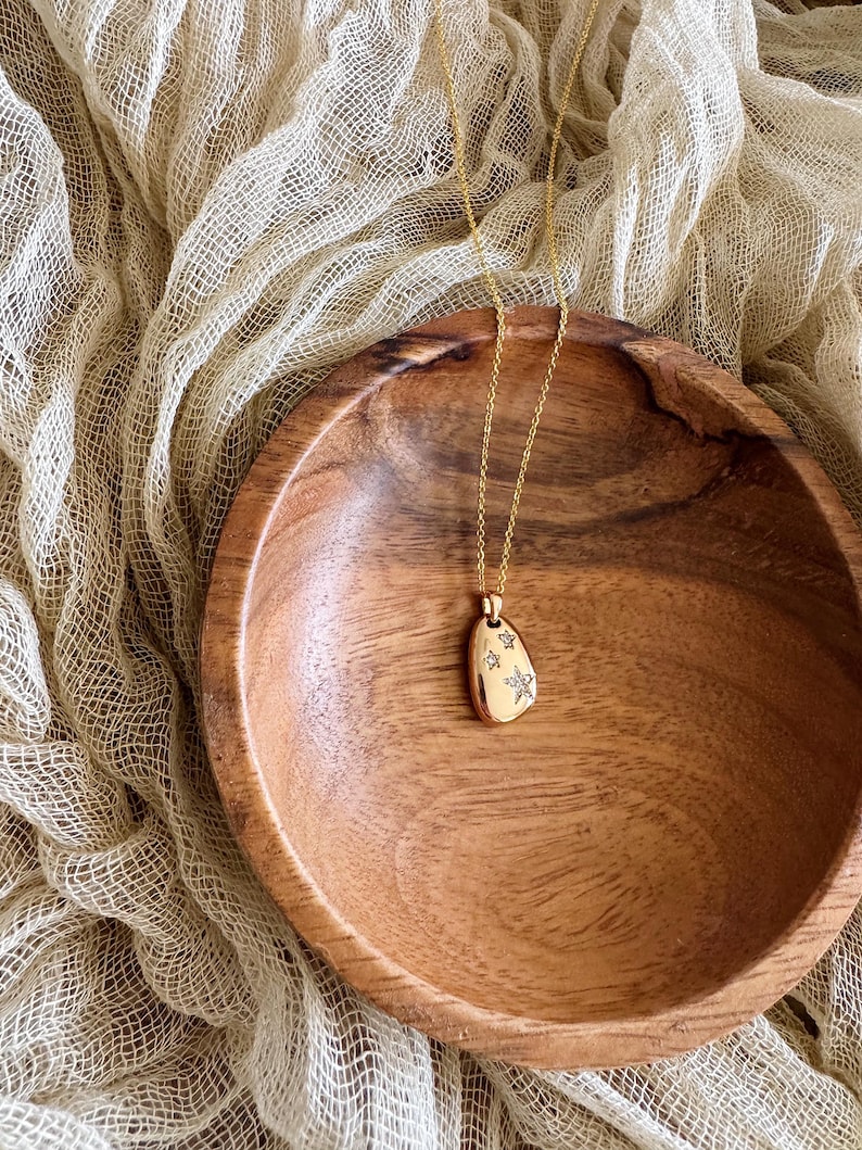 Puede incluir: Un collar de oro con un peque&ntilde;o colgante de forma ovalada que presenta un dise&ntilde;o de estrellas con peque&ntilde;as piedras transparentes. El collar se muestra en un cuenco de madera.