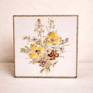 May include: A square ceramic tile featuring a watercolour-style illustration of a wildflower bouquet. The arrangement includes yellow, brown, and muted green floral elements centred on a cream-coloured background with a textured gold-toned border.
