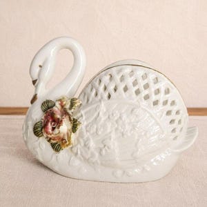 May include: A white ceramic swan figurine featuring a lattice-patterned body and a sculpted, hand-painted rose with green leaves on its chest. The swan has a long, curved neck, a gold-painted beak, and a textured wing design on its side.
