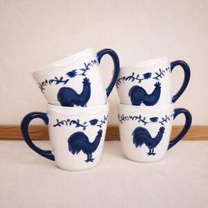 May include: A set of four white ceramic mugs featuring a hand-painted blue rooster motif on the front and a blue vine pattern around the upper rim. Each mug has a solid blue handle and a smooth, rounded body, arranged in a stacked formation.