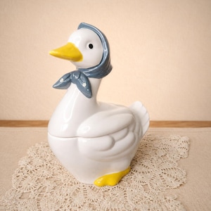 May include: A white ceramic goose figurine with a glossy finish. The goose features a bright yellow beak and feet, and wears a blue headscarf tied under its chin with small white polka dots. The sculpture is shaped as a lidded container.
