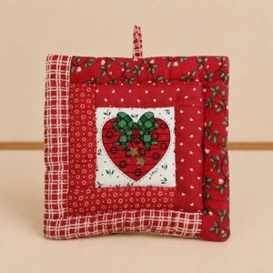 Op de afbeelding: Een vierkante gewatteerde pannenlap met een wit middenpaneel met een rode hartafbeelding, een groen geruite strik en twee kleine gouden sterren. De rand bestaat uit patchworkstof in rood met witte hartjes, rood ruitpatroon en groene bladeren.