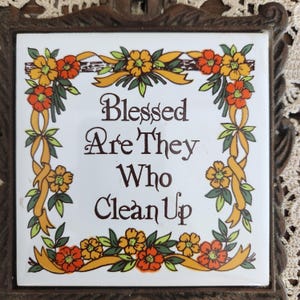 May include: Square ceramic tile with the words "Blessed Are They Who Clean Up" in brown script. The tile is framed with a brown metal border and decorated with orange and yellow flowers and ribbons.