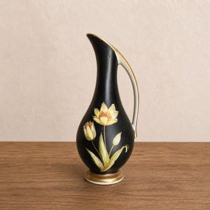 May include: A black ceramic pitcher with a slender neck and a single handle. The body features a painted floral design of yellow tulips with green stems and leaves. The rim, handle, and circular base are accented with a metallic gold-toned finish.
