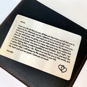 May include: A silver metal wallet card with a heartfelt message, resting on a dark brown leather wallet. The card features a romantic message and two heart symbols. The wallet is a classic accessory.