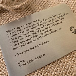 May include: A rectangular brushed metal wallet card featuring an engraved rose graphic and the text: When I say I love you more, I don't mean I love you more than you love me. I mean I love you more than the bad days ahead of us. I love you more than any fight we will ever have. I love you more than the distance between us. I love you more than any obstacle that could ever try and come between us. I love you the most Andy. Love, Your Little Schmoo.