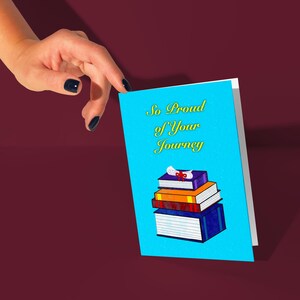May include: A blue card with the text "So Proud of Your Journey" in yellow script. A stack of colorful books with a graduation cap on top is illustrated on the card.