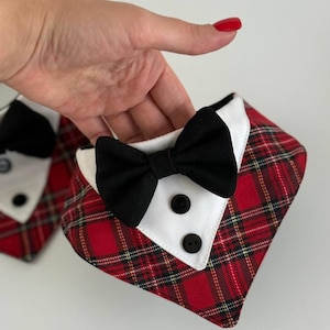 May include: Red plaid dog bandana with a white collar, black bow tie, and two black buttons. The bandana is designed to look like a tuxedo. The image shows two bandanas, one in the foreground and one in the background.