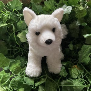May include: A white plush dog toy with a black nose and eyes, set against a backdrop of green leaves. The toy dog has a fluffy texture and is standing upright, showcasing its ears and tail.