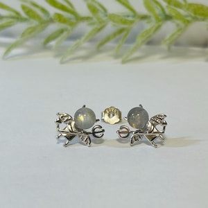 Könnte beinhalten: Ein Paar silberne Ohrringe mit einem kleinen, runden, schillernden Edelstein und einem geflügelten Teufelsdesign.