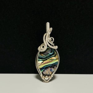 Peut inclure: Pendentif artisanal avec une coquille d'ormeau sertie dans un fil d'argent. La coquille présente des reflets irisés bleus, verts et violets. Le fil forme une bélière décorative et un cadre détaillé autour de la coquille, créant un bijou unique.