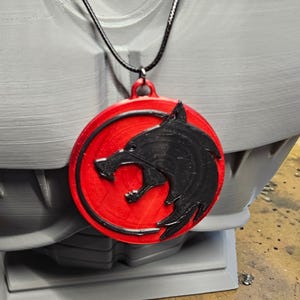 May include: A red and black pendant necklace featuring a wolf head design. The pendant is circular with a black wolf head on a red background. The necklace is displayed on a gray bust.