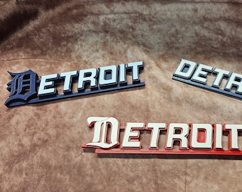May include: Three different versions of the word "DETROIT" in white letters with a black outline. The first version is in a bold, blocky font. The second version is in a more traditional font. The third version is in a stylized, gothic font. The first version is on a dark blue background, the second version is on a light blue background, and the third version is on a red background.