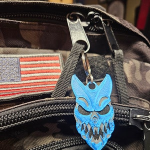 May include: A blue and black skull keychain with sharp teeth and a menacing expression. The keychain is attached to a black zipper pull on a camouflage backpack.
