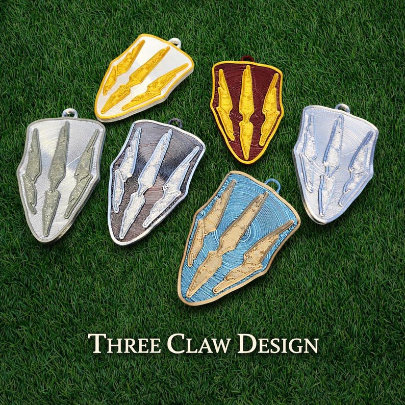 Puede incluir: Varios colgantes en forma de escudo con un dise&ntilde;o de tres garras. Los colgantes son de color oro, plata y azul, y est&aacute;n dispuestos sobre una superficie verde. El texto "THREE CLAW DESIGN" es visible en la parte inferior.