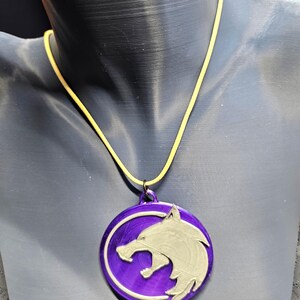 Puede incluir: Un collar con colgante morado con un dise&ntilde;o de cabeza de lobo plateado. El colgante circular cuelga de un cord&oacute;n amarillo. El collar se exhibe en un maniqu&iacute;.