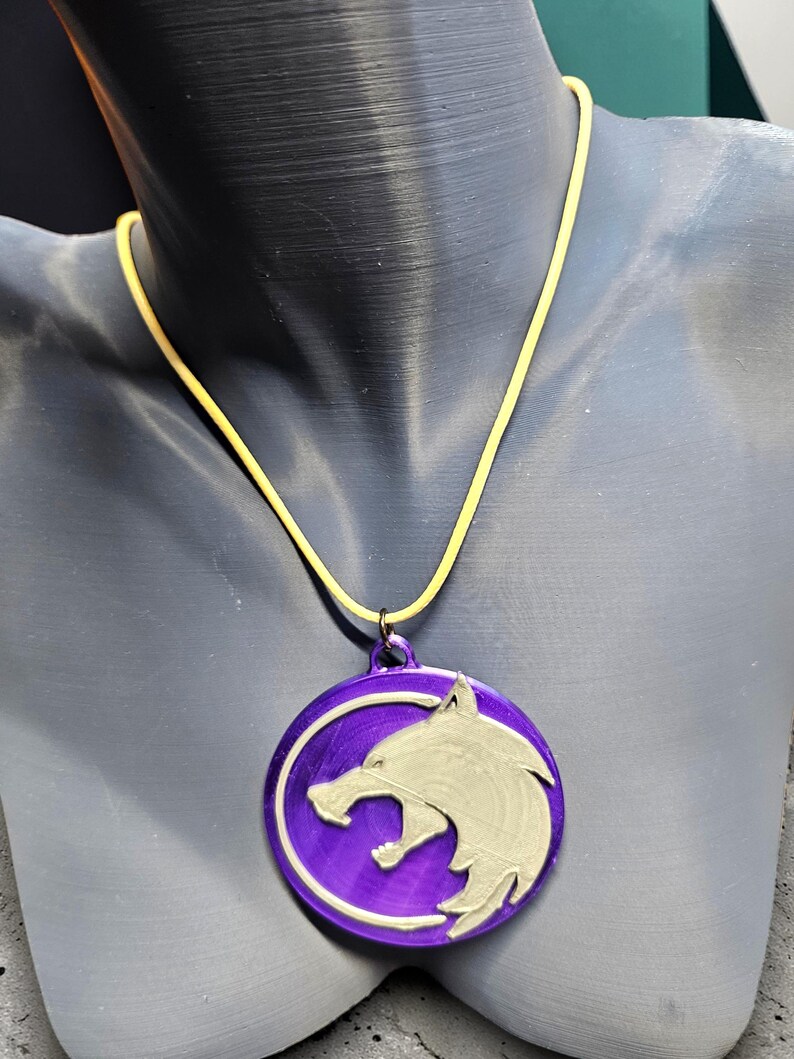 Puede incluir: Un collar con colgante morado con un dise&ntilde;o de cabeza de lobo plateado. El colgante circular cuelga de un cord&oacute;n amarillo. El collar se exhibe en un maniqu&iacute;.