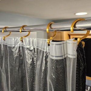 May include: A transparent shower curtain with a dark pattern suspended from a silver rail with gold-coloured hooks. The curtain allows light to pass through. The curved hooks secure the curtain. The shower curtain is in a bathroom.