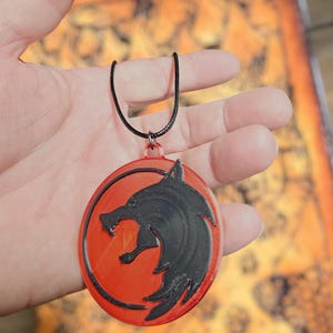 Puede incluir: Un collar con colgante redondo con un dise&ntilde;o de cabeza de lobo negro sobre fondo rojo. El colgante cuelga de un cord&oacute;n negro. Un accesorio tem&aacute;tico.