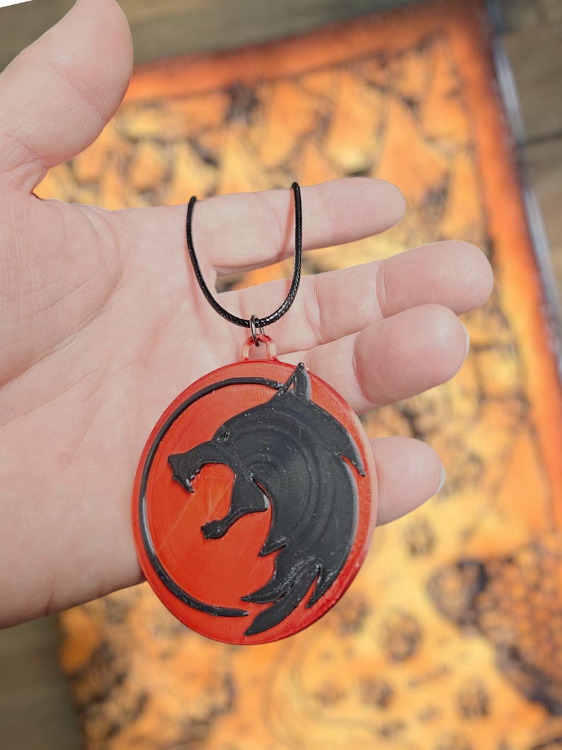 Puede incluir: Un collar con colgante redondo con un dise&ntilde;o de cabeza de lobo negro sobre fondo rojo. El colgante cuelga de un cord&oacute;n negro. Un accesorio tem&aacute;tico.