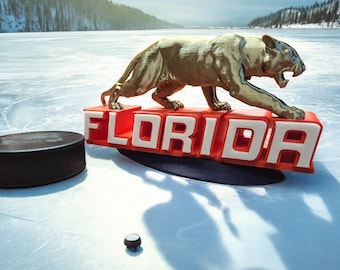 Puede incluir: Una estatua dorada de un puma sobre un letrero rojo y blanco con la palabra "FLORIDA", con un disco de hockey y un pequeño objeto negro sobre una superficie helada. El fondo presenta un paisaje nevado con árboles y un lago.