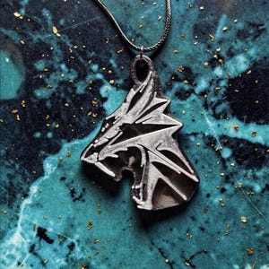 Puede incluir: Un collar con colgante plateado que representa una cabeza de lobo estilizada. El colgante cuelga de una fina cadena plateada. La cabeza de lobo tiene rasgos afilados y angulares. El fondo es una superficie veteada en azul turquesa y azul oscuro con motas doradas.