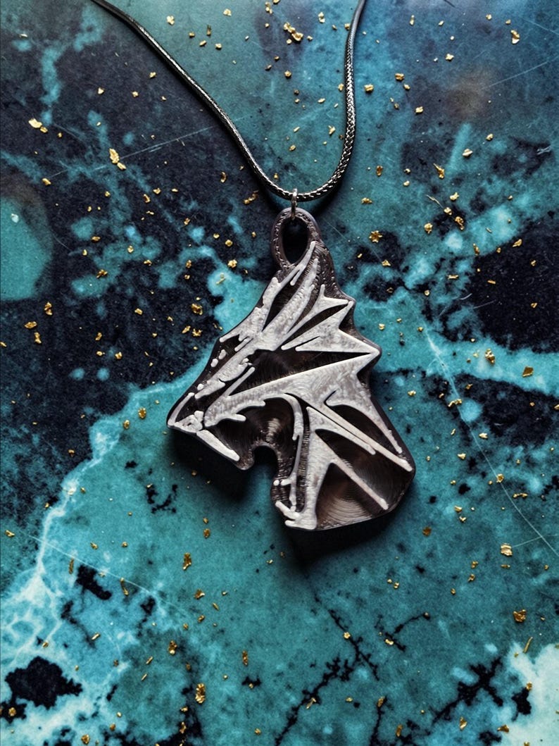 Puede incluir: Un collar con colgante plateado que representa una cabeza de lobo estilizada. El colgante cuelga de una fina cadena plateada. La cabeza de lobo tiene rasgos afilados y angulares. El fondo es una superficie veteada en azul turquesa y azul oscuro con motas doradas.