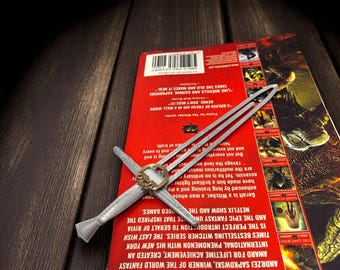 Puede incluir: Una espada plateada en miniatura descansa sobre un libro rojo. La portada del libro presenta texto e imágenes, incluyendo "The Last Wish" y "Andrzej Sapkowski". La espada tiene una empuñadura y guarda detalladas.
