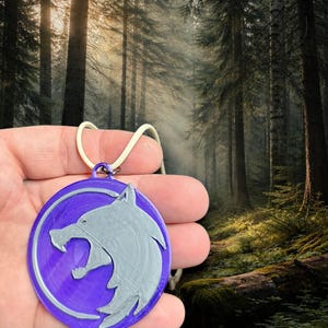 Puede incluir: Un colgante morado circular con un dise&ntilde;o de lobo plateado, suspendido de un cord&oacute;n blanco. El colgante presenta una cabeza de lobo en relieve. El fondo muestra una escena de bosque con &aacute;rboles altos y luz solar.