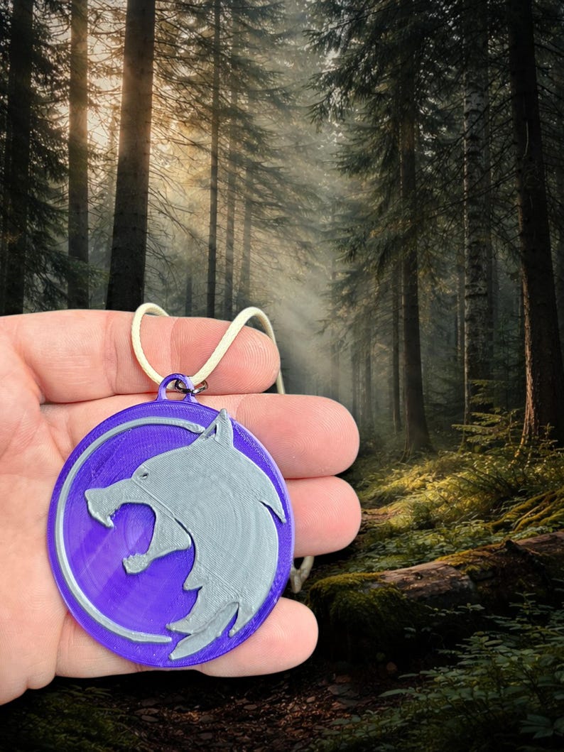 Puede incluir: Un colgante morado circular con un dise&ntilde;o de lobo plateado, suspendido de un cord&oacute;n blanco. El colgante presenta una cabeza de lobo en relieve. El fondo muestra una escena de bosque con &aacute;rboles altos y luz solar.