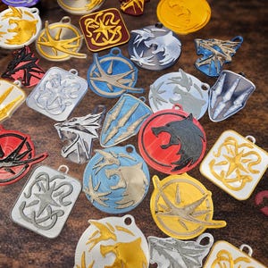 Puede incluir: Una colecci&oacute;n de colgantes coloridos impresos en 3D con varios dise&ntilde;os de fantas&iacute;a. Los colgantes presentan emblemas de lobos, p&aacute;jaros y otros estilizados en oro, plata, rojo, azul y negro. Cada colgante tiene un peque&ntilde;o lazo.