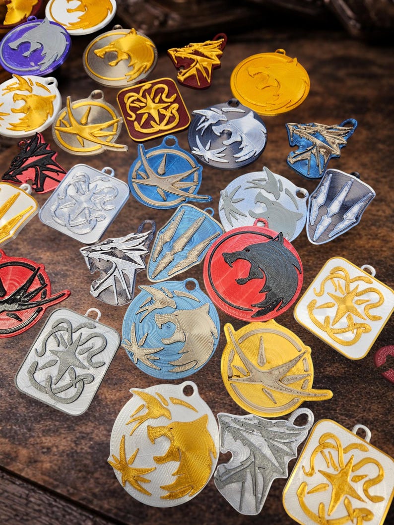 Puede incluir: Una colecci&oacute;n de colgantes coloridos impresos en 3D con varios dise&ntilde;os de fantas&iacute;a. Los colgantes presentan emblemas de lobos, p&aacute;jaros y otros estilizados en oro, plata, rojo, azul y negro. Cada colgante tiene un peque&ntilde;o lazo.