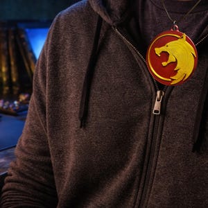 Puede incluir: Un collar con un colgante redondo rojo y amarillo con un dise&ntilde;o de lobo. El colgante cuelga de una fina cadena dorada. La persona lleva una sudadera con capucha gris oscuro con cremallera.