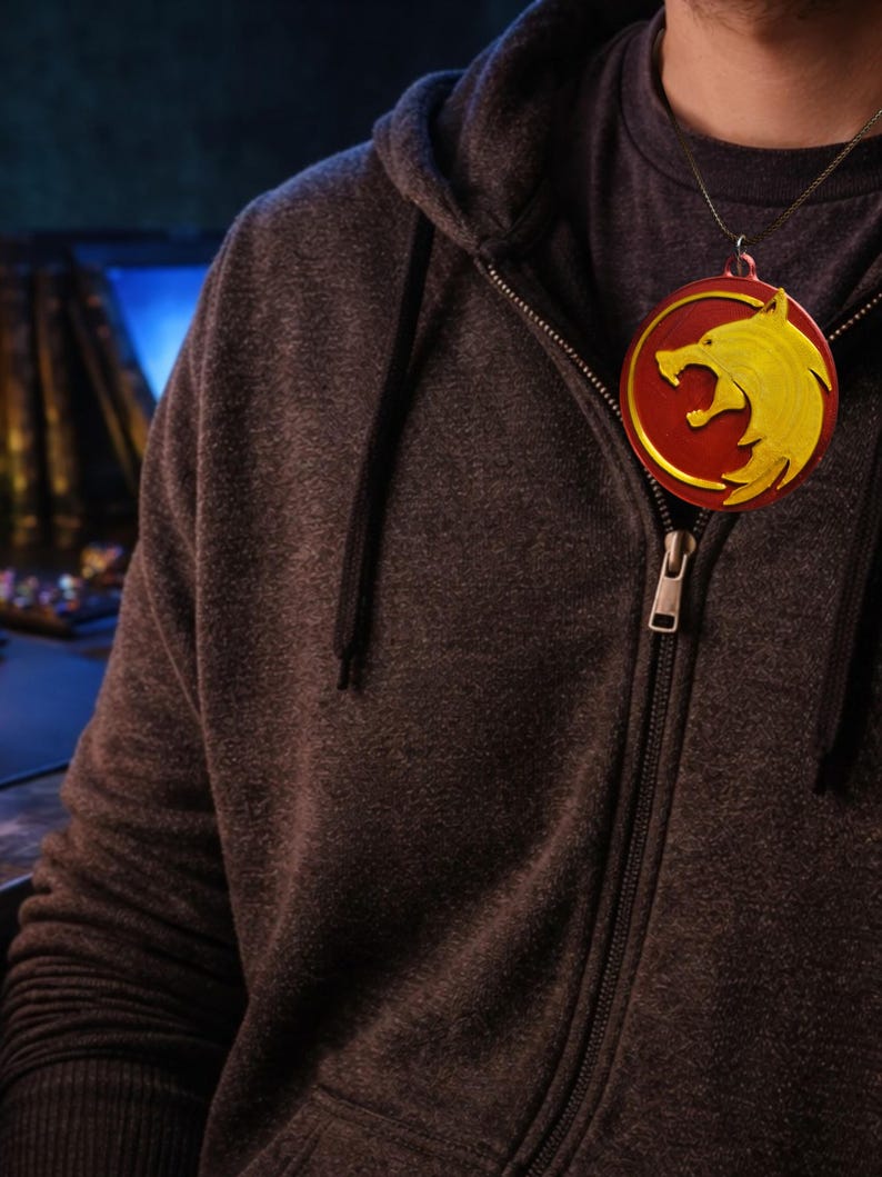 Puede incluir: Un collar con un colgante redondo rojo y amarillo con un dise&ntilde;o de lobo. El colgante cuelga de una fina cadena dorada. La persona lleva una sudadera con capucha gris oscuro con cremallera.