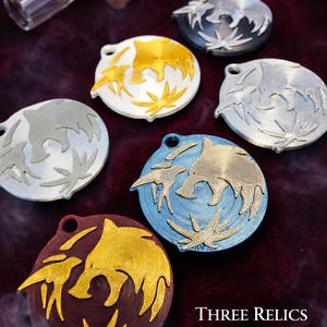 Puede incluir: Varios colgantes circulares met&aacute;licos de diferentes colores, con un dise&ntilde;o estilizado de animal. Se muestran sobre un fondo oscuro y texturizado, con una peque&ntilde;a botella de vidrio y un corcho. Tambi&eacute;n se ve el texto "THREE RELICS DESIGN".