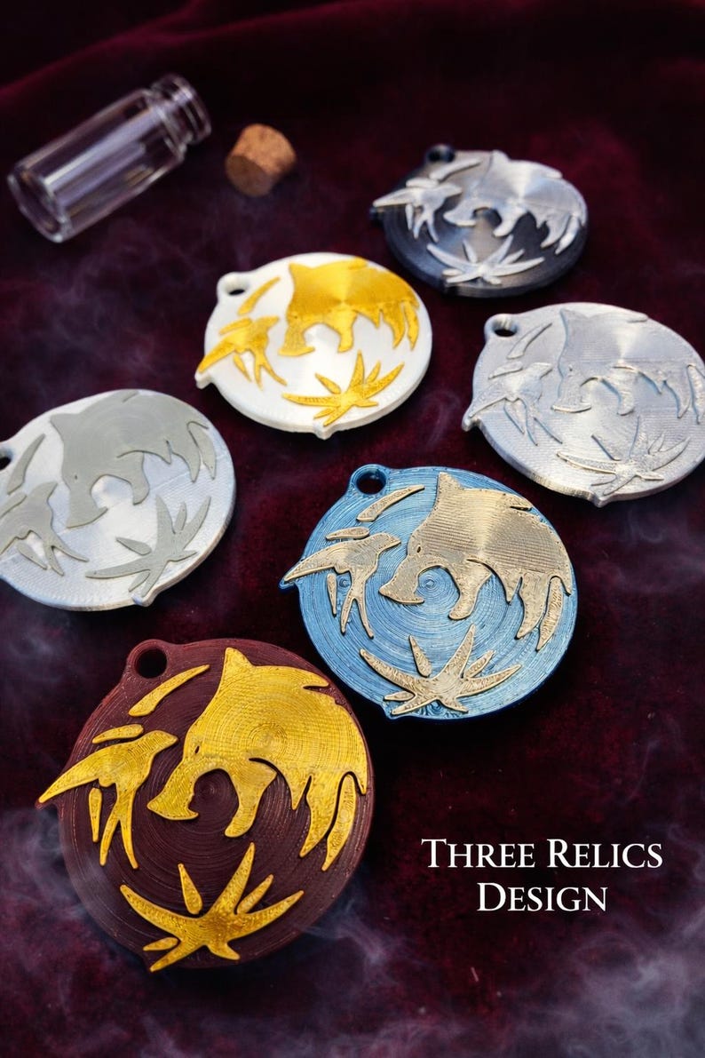 Puede incluir: Varios colgantes circulares met&aacute;licos de diferentes colores, con un dise&ntilde;o estilizado de animal. Se muestran sobre un fondo oscuro y texturizado, con una peque&ntilde;a botella de vidrio y un corcho. Tambi&eacute;n se ve el texto "THREE RELICS DESIGN".