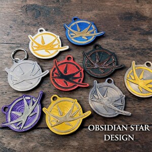 Puede incluir: Una colecci&oacute;n de llaveros y colgantes Obsidian Star Design de colores. Los dise&ntilde;os presentan un emblema circular con un dise&ntilde;o de estrella estilizado en varios colores, incluyendo blanco, amarillo, rojo, azul y plateado. El texto "OBSIDIAN STAR DESIGN" es visible.