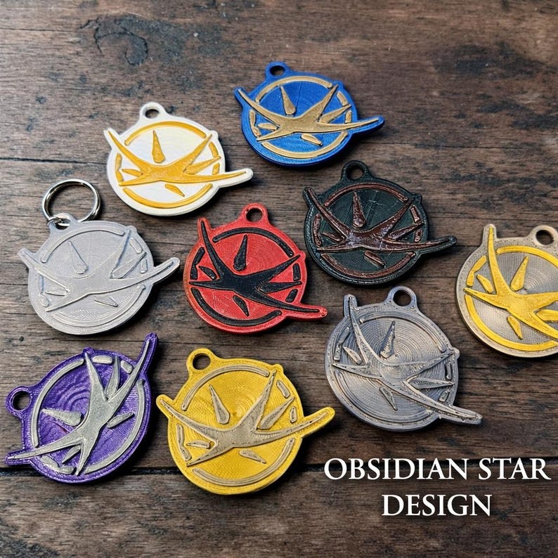 Puede incluir: Una colecci&oacute;n de llaveros y colgantes Obsidian Star Design de colores. Los dise&ntilde;os presentan un emblema circular con un dise&ntilde;o de estrella estilizado en varios colores, incluyendo blanco, amarillo, rojo, azul y plateado. El texto "OBSIDIAN STAR DESIGN" es visible.