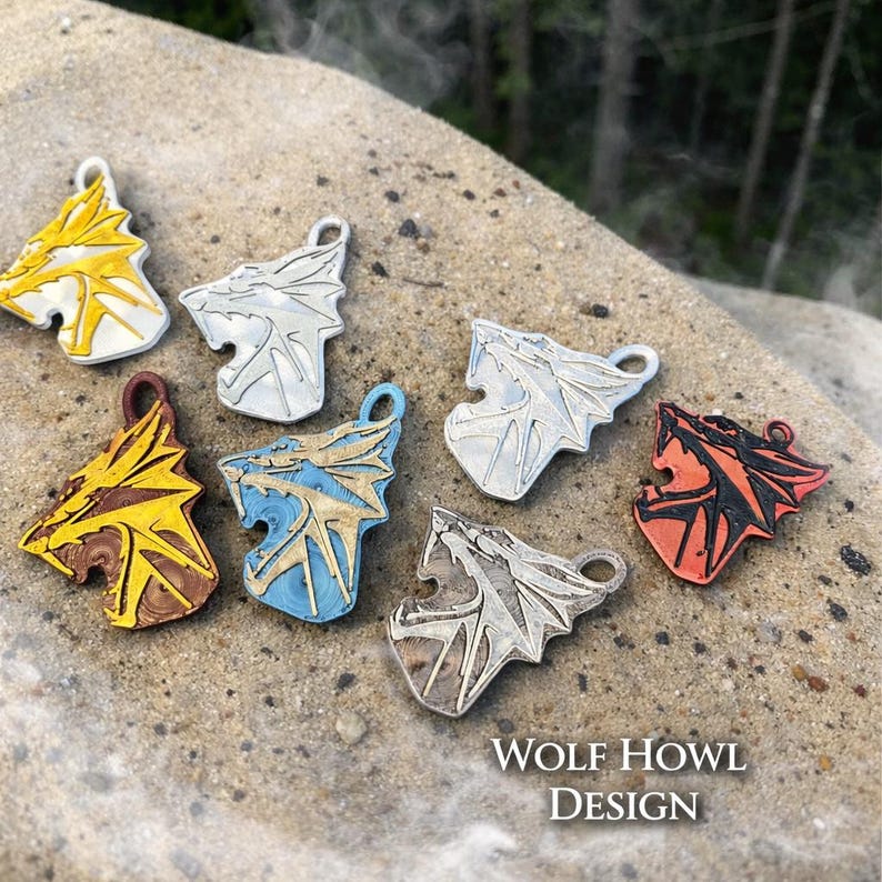 Puede incluir: Varios colgantes de cabeza de lobo en oro, plata, azul y rojo. Cada colgante presenta un dise&ntilde;o detallado de un lobo aullando. El texto "WOLF HOWL DESIGN" est&aacute; en la parte inferior.