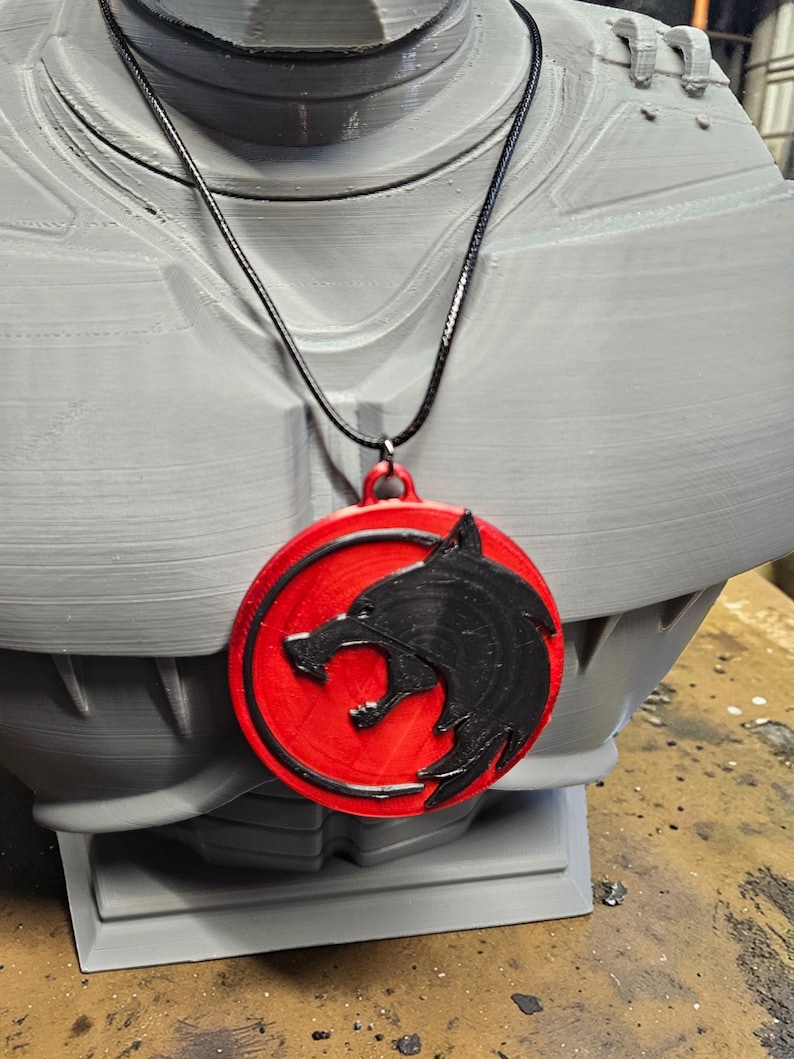 May include: A red and black pendant necklace featuring a wolf head design. The pendant is circular with a black wolf head on a red background. The necklace is displayed on a gray bust.