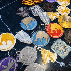 Puede incluir: Una colecci&oacute;n de llaveros impresos en 3D de varias formas y colores. Los llaveros presentan dise&ntilde;os de animales y s&iacute;mbolos, incluyendo jabal&iacute;es, escudos y emblemas estilizados. La paleta de colores incluye dorado, plateado, azul, rojo y morado.