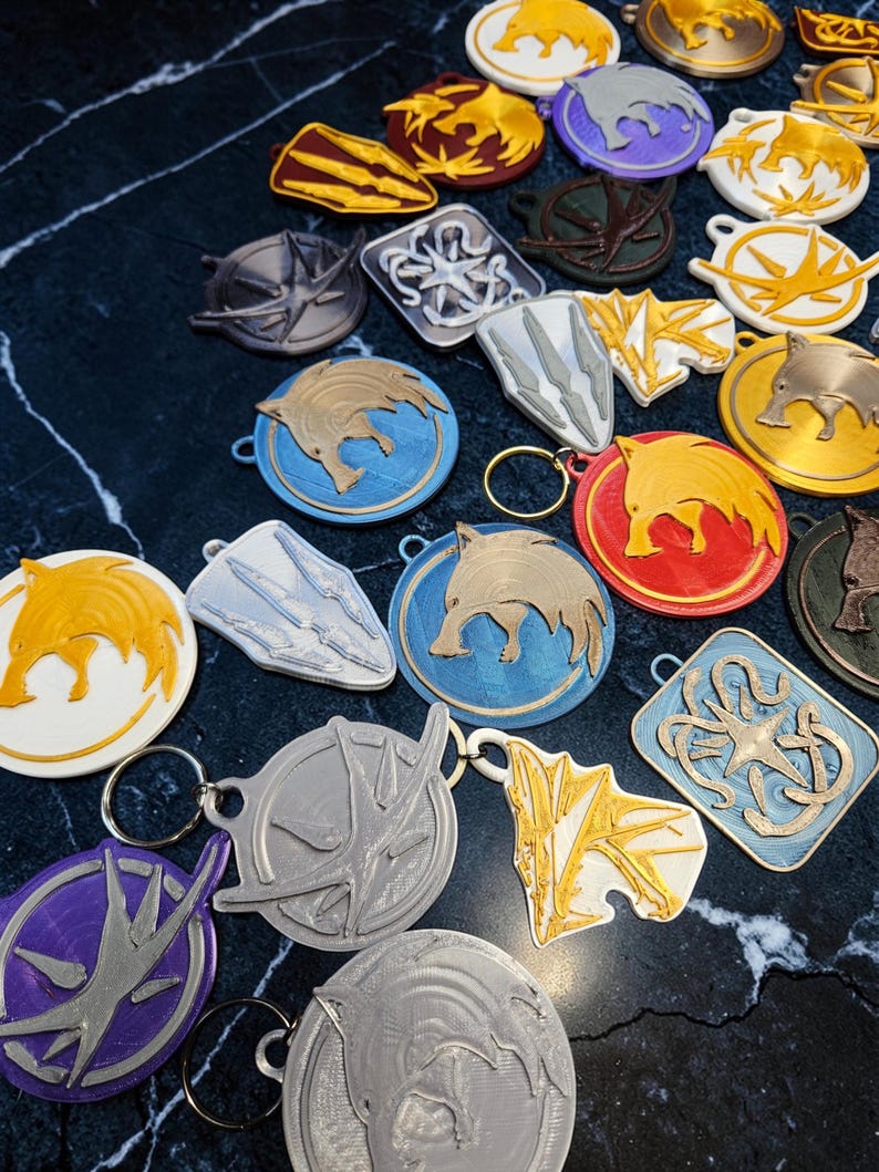 Puede incluir: Una colecci&oacute;n de llaveros impresos en 3D de varias formas y colores. Los llaveros presentan dise&ntilde;os de animales y s&iacute;mbolos, incluyendo jabal&iacute;es, escudos y emblemas estilizados. La paleta de colores incluye dorado, plateado, azul, rojo y morado.