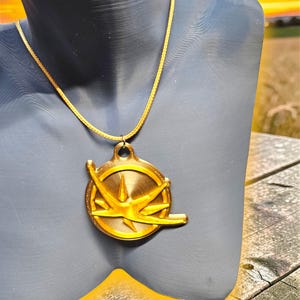 Puede incluir: Un collar con colgante dorado. El colgante circular presenta un dise&ntilde;o de estrella y un lazo en la parte superior. La cadena tambi&eacute;n es dorada. El collar se muestra en un maniqu&iacute; gris.