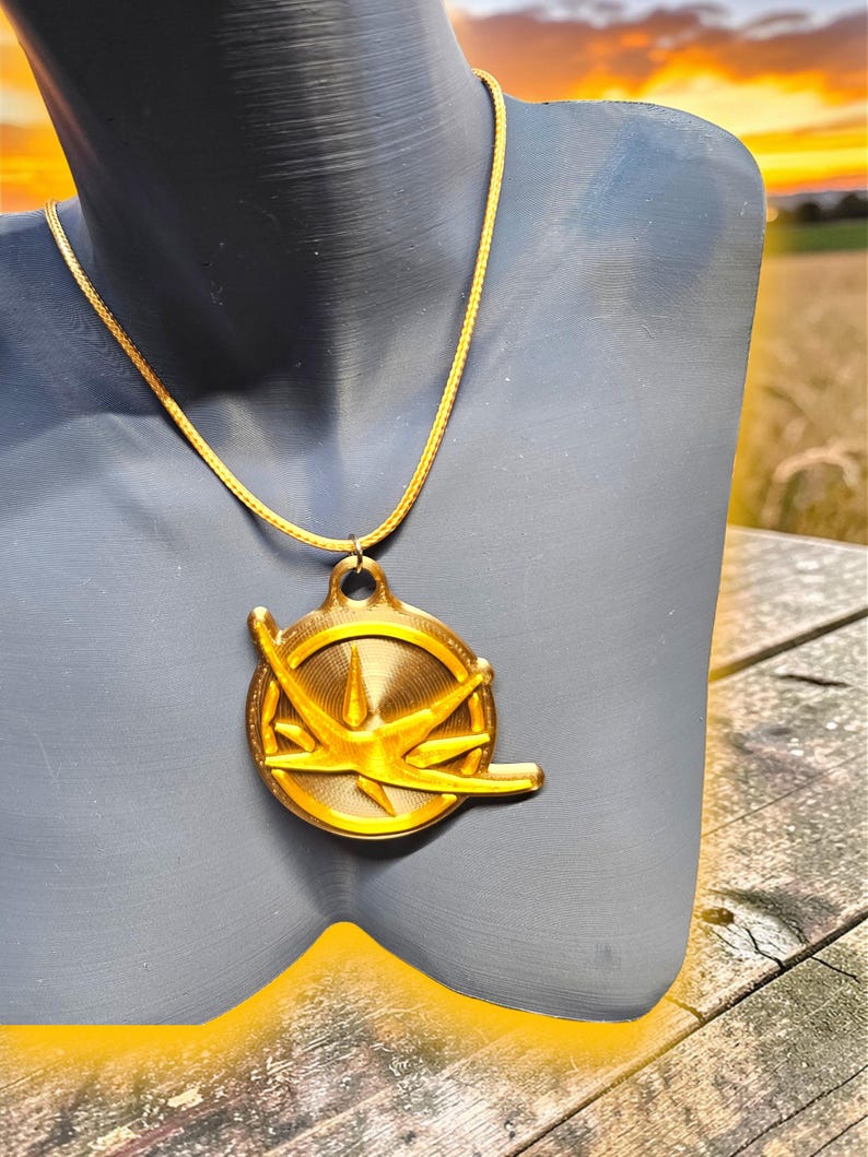 Puede incluir: Un collar con colgante dorado. El colgante circular presenta un dise&ntilde;o de estrella y un lazo en la parte superior. La cadena tambi&eacute;n es dorada. El collar se muestra en un maniqu&iacute; gris.