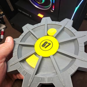 May include: A gray, gear-shaped object with a yellow center and accents. The center circle has the number "67" in black. The object is held in a person's hand, with a computer in the background.