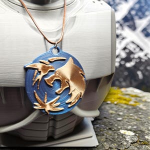 Puede incluir: Un collar con colgante azul y dorado con un dise&ntilde;o de lobo. El colgante ovalado presenta una cabeza de lobo dorada y detalles dorados sobre un fondo azul. El collar se muestra sobre un busto blanco, con monta&ntilde;as al fondo.