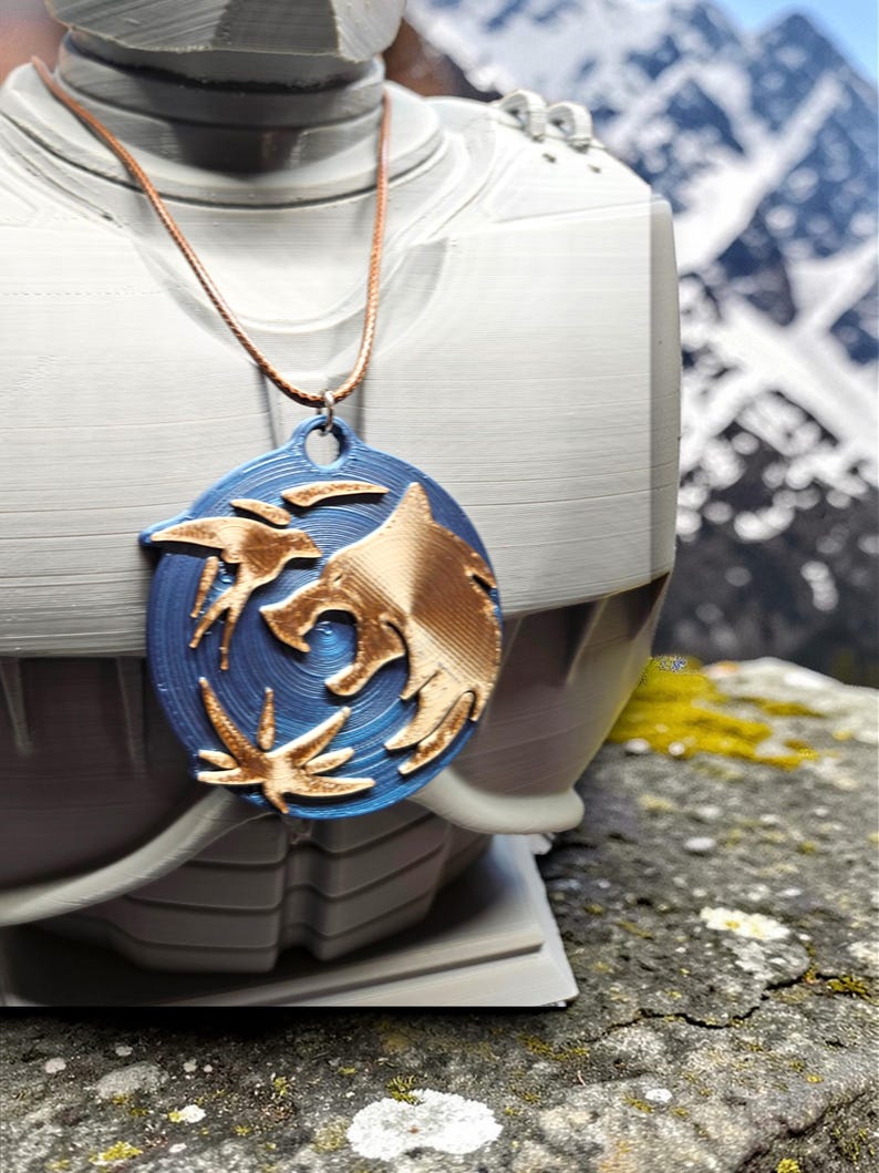 Puede incluir: Un collar con colgante azul y dorado con un dise&ntilde;o de lobo. El colgante ovalado presenta una cabeza de lobo dorada y detalles dorados sobre un fondo azul. El collar se muestra sobre un busto blanco, con monta&ntilde;as al fondo.