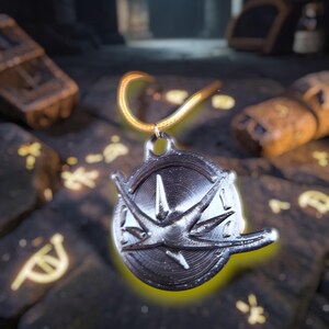 Puede incluir: Un colgante plateado con un dise&ntilde;o de rosa de los vientos, suspendido de una cadena dorada. El colgante es circular con un patr&oacute;n de estrella en relieve. El fondo es una superficie oscura, similar a la piedra, con s&iacute;mbolos brillantes y un entorno cavernoso borroso.