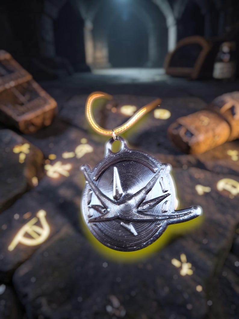 Puede incluir: Un colgante plateado con un dise&ntilde;o de rosa de los vientos, suspendido de una cadena dorada. El colgante es circular con un patr&oacute;n de estrella en relieve. El fondo es una superficie oscura, similar a la piedra, con s&iacute;mbolos brillantes y un entorno cavernoso borroso.